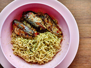 A simple budget-friendly meal of instant noodles topped with spicy canned fish, served in a pink bowl on a wooden table. Popular comfort food and an easy homemade dish in many Asian households.