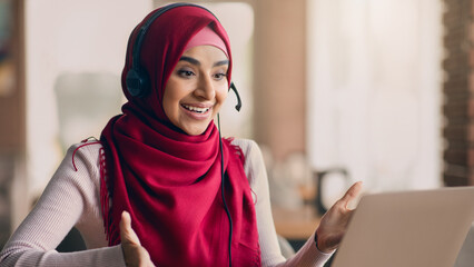 A young woman engages in an online learning session, smiling and speaking into a headset. She wears a bright red hijab and sits at a desk in a warm, inviting space with natural light.
