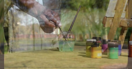 Mixing mature man wearing striped apron stirring paintbrushes at wood table in garden, paint jars