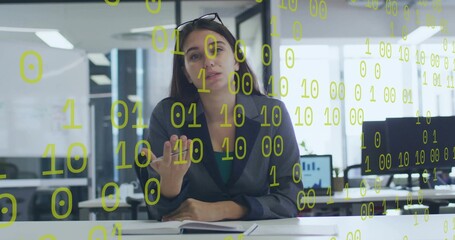 Presenting woman in dark blazer, teal top gesturing at desk in office, notebook, binary overlay