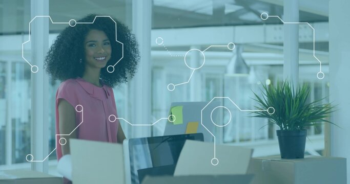 Smiling woman wearing pink blouse standing at desk in modern office with laptop monitor boxes plant