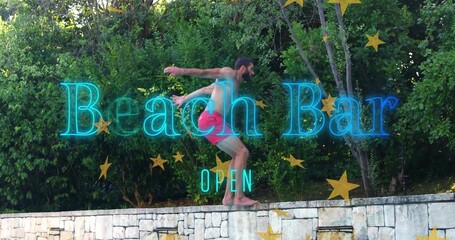 Crouching man in red trunks balancing on stone wall in backyard with neon Beach Bar OPEN