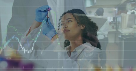 Scientist wearing lab coat and blue nitrile gloves holding vial against light in lab with charts