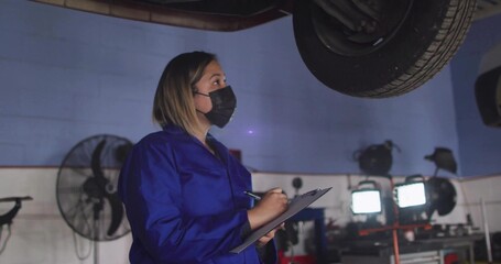 Inspecting woman in blue coveralls wearing black mask with clipboard and pen under lifted car