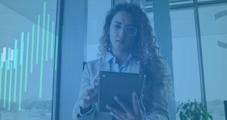 Standing woman checking tablet by large windows with teal chart overlay, in blazer and glasses