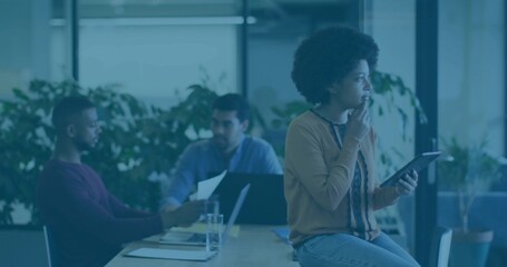 Holding tablet, woman in patterned blouse, jeans gazing out window at table with laptop, papers