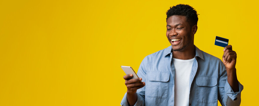 A cheerful man smiles while checking his mobile phone and holding a credit card. The bright yellow background adds energy to the scene, suggesting a moment of excitement or success. - Powered by Adobe