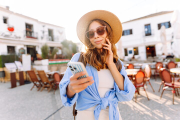 A beautiful woman in a hat stands in a busy square, holding a phone. A young tourist in sunglasses...