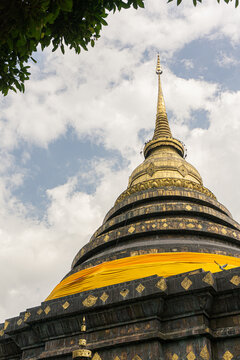 tiered Buddhist stupa (chedi) featuring a dark, heavily adorned base and a conical golden upper section that tapers to a spire, all set against a cloudy sky. The structure is distinctive for the vibra