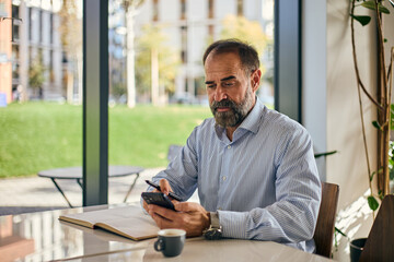 Businessman At Outdoor Caf&eacute; Using Smartphone With Notebook And Coffee In Bright Modern Setting