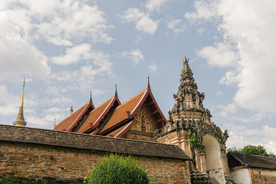 layered architecture of Wat Phra That Lampang Luang, featuring the low, weathered brick wall and the ancient Pratu Khong (Lanna gateway) in the foreground, contrasted with the newer, vibrant red roof 