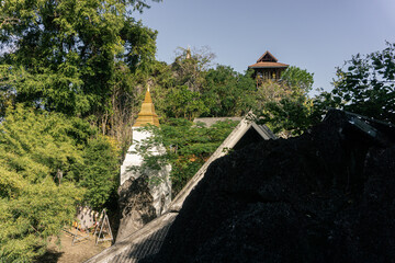 serene Buddhist temple complex, featuring a white and gold stupa partially concealed by dense foliage and rugged rocks, emphasizing the integration of spiritual structure with the wild, natural enviro