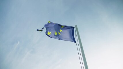 A tattered European Union flag fluttering in the wind against blue sky symbolizing hardship and endurance. Low angle view.