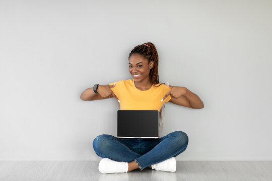 Pretty young black woman sits cross legged on the floor, pointing joyfully at an empty laptop displaying a website design mockup in a grey studio space during daylight.