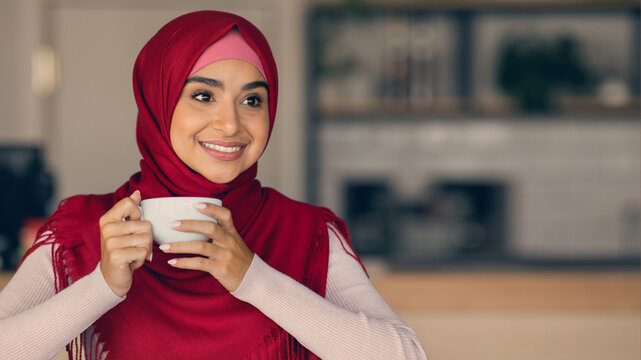 A young woman wearing a hijab holds a cup of coffee while smiling in a bright and inviting cafe. The interior has a warm ambiance with soft lighting and minimal decor. - Powered by Adobe