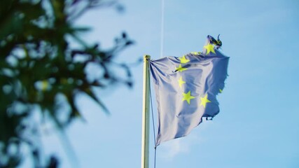 A tattered European Union flag fluttering in the wind against blue sky symbolizing hardship and endurance. Low angle view.