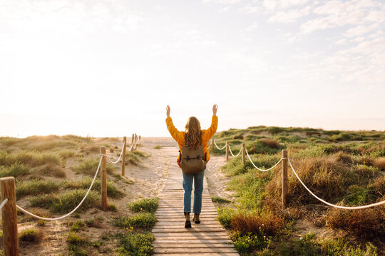 Beautiful woman in bright jacket walks along wooden path overlooking sea. Backpacker enjoys the sunset and feels the freedom of the outdoors. Concepts of hiking, enjoyment, and nature.
