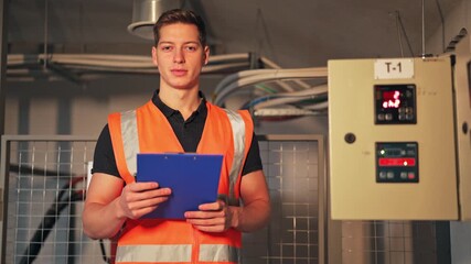 Industrial worker, wearing an orange safety vest, confidently holds a clipboard near electrical panels. Professional and reliable, conveys satisfaction and competence within the technical environment.