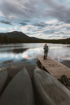 Woman stands on dock with canoes on shore of lake in Maine