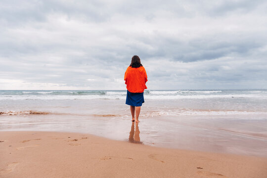 Woman standing barefoot on sandy beach facing the sea under dramatic sky