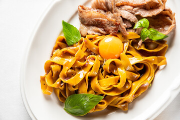 Gourmet Italian tagliatelle with seared meat strips, a vibrant raw egg yolk center, and fresh basil garnish, served on a white plate in a restaurant setting