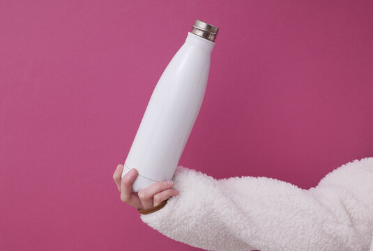 White reusable metal thermos bottle in a female hand wearing a sherpa sweatshirt on a pink background. Mockup for design
