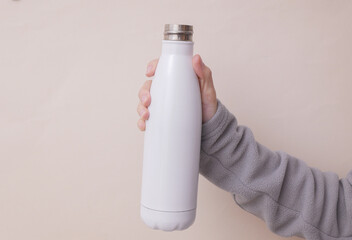 Hand in sports fleece sweatshirt holds white metal reusable bottle. Woman holds thermoson light beige background. Mockup for design. © VI-stock studio