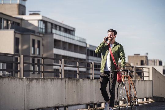 Smiling man talking on smart phone and walking with bicycle near railing