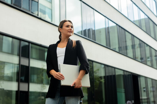 Businesswoman walking outdoors with laptop near modern office building - Powered by Adobe