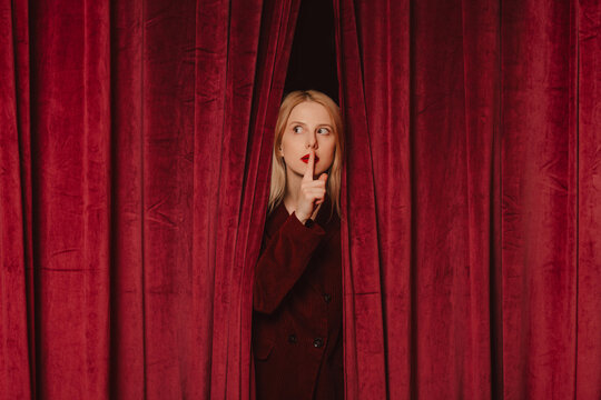 Elegant woman in velvet coat making quiet gesture behind red curtains