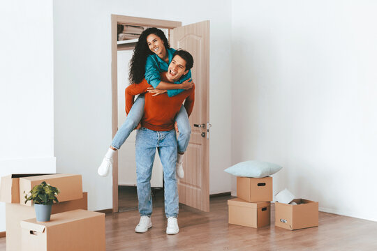 A man carries a woman on his back as they move into their new apartment. They smile happily in a room filled with cardboard boxes and a small plant. The atmosphere is joyful and lively. - Powered by Adobe