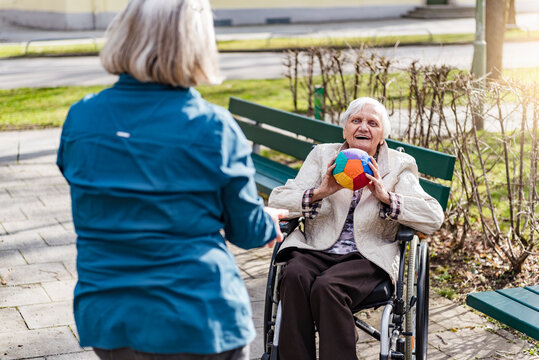 Woman taking care of old woman in wheelchair playing with ball in park