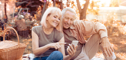 On a warm sunny afternoon, a couple sits close in a park, sharing a light moment as they look at a smartphone together. They are happy and relaxed, surrounded by nature.