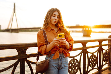 A portrait of a young woman with a phone stands on the embankment at sunset. A beautiful woman texts on her phone and enjoys a walk outdoors. Concept: technology, urban, blogging. Lifestyle.