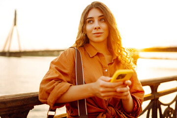 A portrait of a young woman with a phone stands on the embankment at sunset. A beautiful woman...