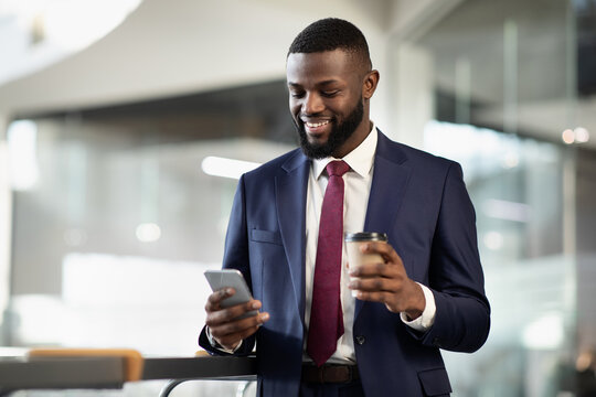 Cheerful young black bearded man manager standing at office building corridor, drinking coffee to go, using mobile phone, chatting with business partner and smiling, panorama with copy space