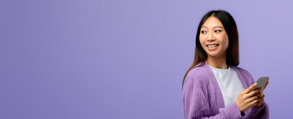 A woman stands against a soft purple background, holding her smartphone while smiling to the side....