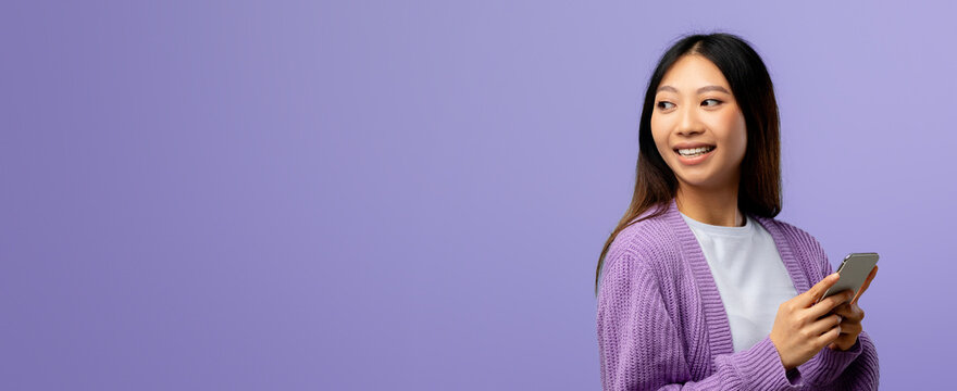 A woman stands against a soft purple background, holding her smartphone while smiling to the side. She wears a cozy sweater, embodying a relaxed atmosphere. - Powered by Adobe