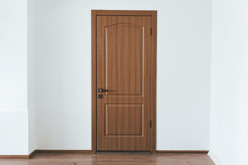 A wooden door stands closed in a bright room. The door features a classic design with a smooth finish. The surrounding walls are painted white, enhancing the spacious feel.