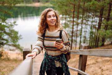 Portrait of a traveler with a backpack and a phone stands on a wooden path by a lake. A sweet woman...