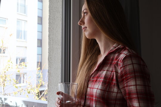 A thoughtful and smiling young woman is standing in front of a window and holding a glass of water in her hands.