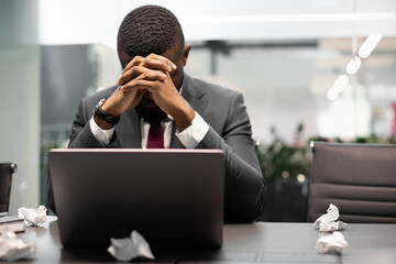 Closeup portrait of tired black manager suffering from burnout, sitting at table and working on laptop at office, leaning on his hands, looking for creative ideas for business, copy space