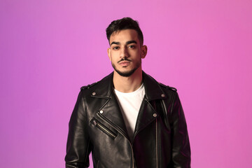 A handsome young Arab man stands confidently, wearing a black leather jacket. He looks directly at the camera, against a vibrant neon background, showcasing modern fashion and style.