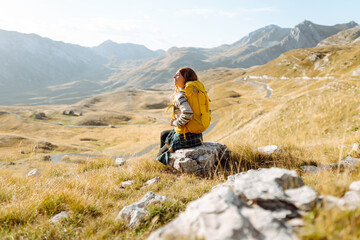 Fototapeta premium Female hiker with backpack stands on mountain trail at sunset. The beautiful woman feels free and enjoys the mountain scenery. Concept of trail, freedom, travel.