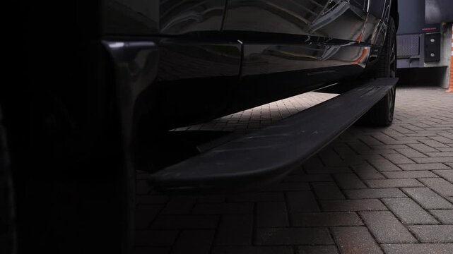 Low angle close up pans along a Range Rover Autobiography as the powered running board extends on a herringbone forecourt near a loading dock cone and ramp in overcast light.