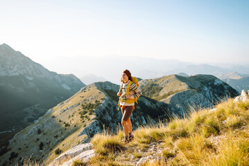 Fototapeta premium Beautiful female hiker with yellow backpack stands on mountain ridge, watching sunset. Young woman enjoys journey and feels freedom of sunset. Concept of hiking, freedom, and an active lifestyle.
