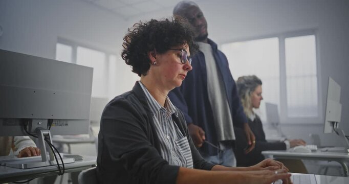 Attentive African American Instructor Walks Between Adult Students During Programming Workshop in Modern Computer Classroom. Helps Debug Code, Provides Feedback, Explains Complex Concepts. Dolly Shot.