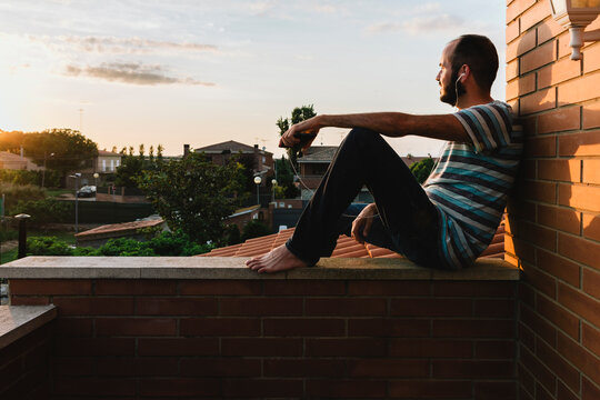 Thoughtful man listening music while sitting on retaining wall against sky during sunset