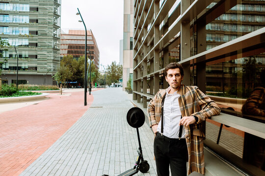 Portrait of stylish man in check jacket with push scooter standing at modern building in city