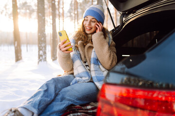 Young woman sits in trunk of car with phone, enjoying a snowy day. A smiling woman texts on her phone and relaxes during a road trip on a sunny winter day. Travel concept. Active lifestyle.